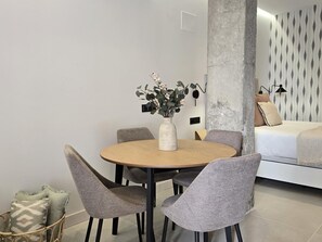 A modern living room with a round wooden table, grey upholstered chairs, a white vase with flowers, and a bed with patterned wallpaper. - Design studios in Pedregalejo - 4 guests (Málaga)