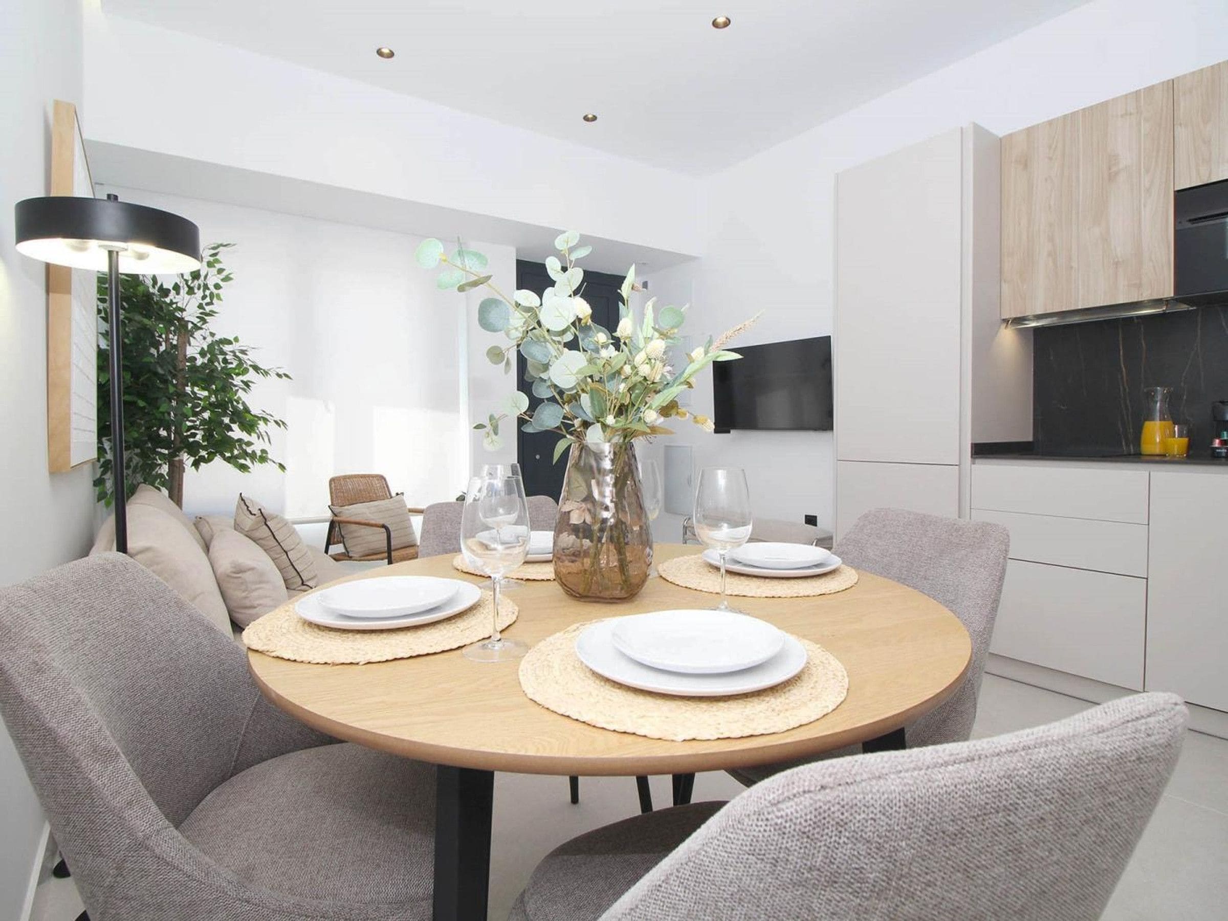 A modern dining area with a round table, grey upholstered chairs, a vase with flowers, and a television mounted on the wall.