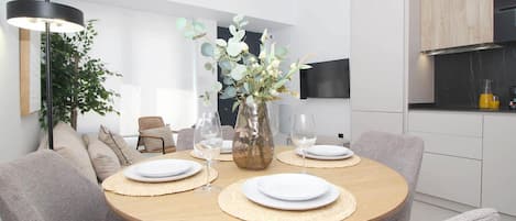 A modern dining area with a round table, grey upholstered chairs, a vase with flowers, and a television mounted on the wall.