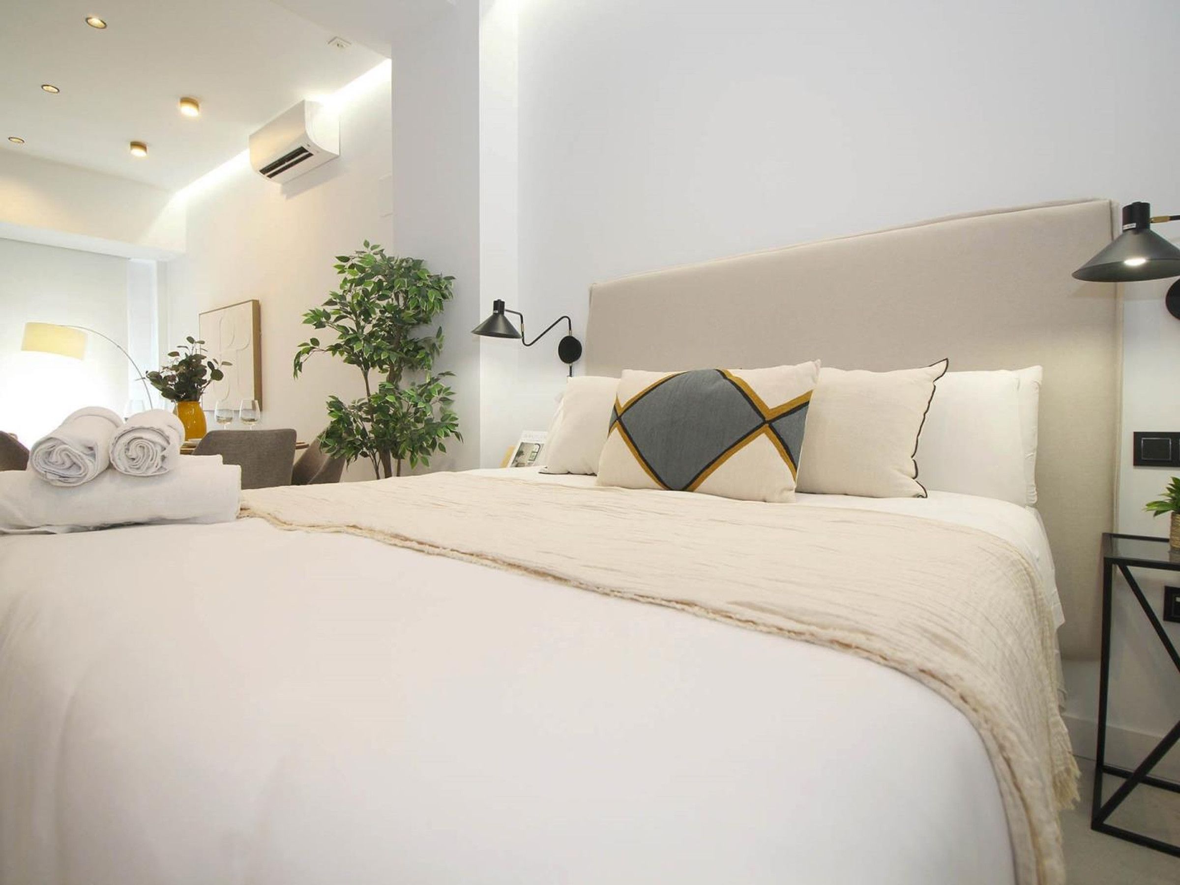 A hotel room with a large bed, a bedside table with a lamp, a plant, and a wall-mounted air conditioner.