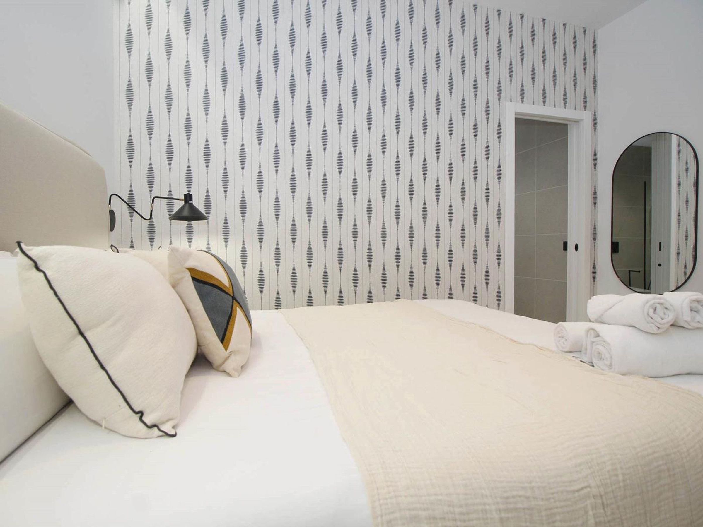 Une chambre d’hôtel comprenant un lit, des oreillers, une lampe de chevet, un miroir et une porte donnant accès à une salle de bain.