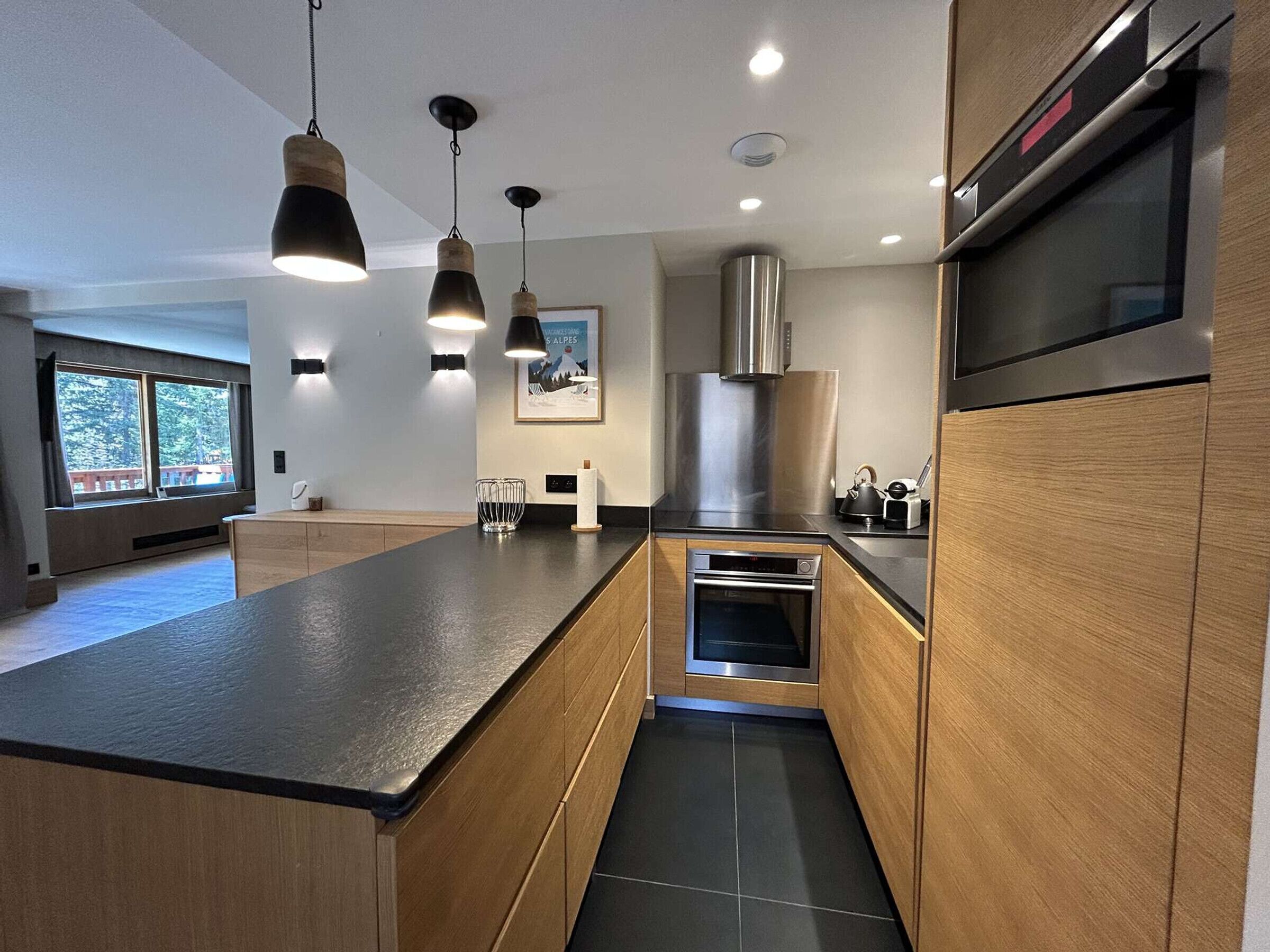 A modern kitchen with a central island, stainless steel appliances, pendant lighting, and a large window providing natural light.