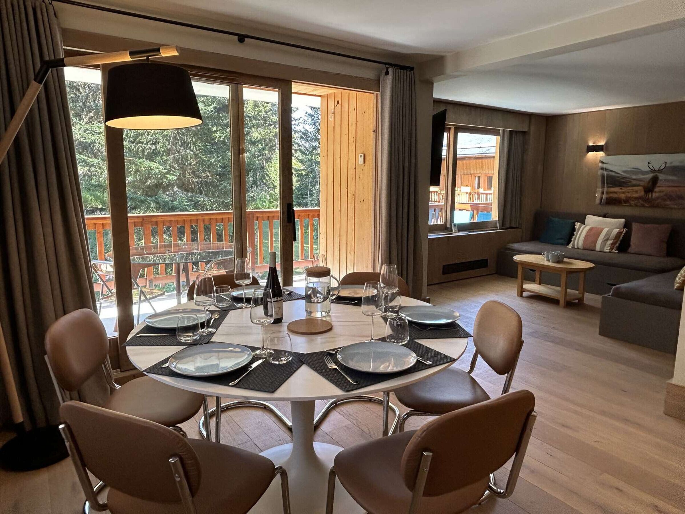 Comedor con una mesa redonda dispuesta para cuatro personas, rodeada de sillas, y vistas a una terraza de madera y árboles al exterior.