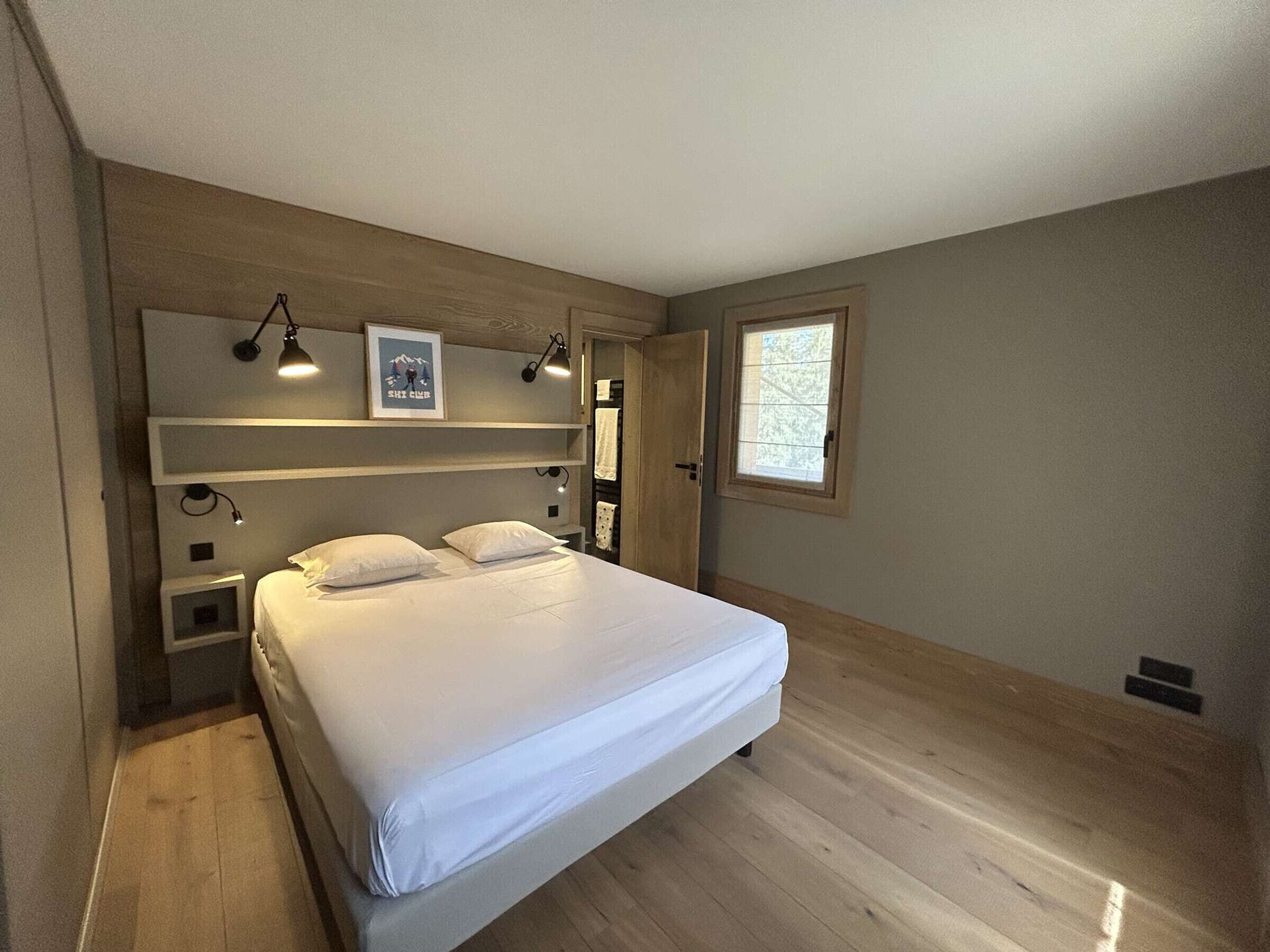 Un dormitorio con cama, mesitas de noche, una ventana e iluminación en la pared.