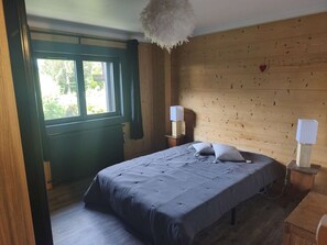 A bedroom with a wooden wall, a bed with a grey bedspread, two bedside tables with lamps, and a window with curtains. - Spacious apartment near center - 8 guests (Arâches-la-Frasse)