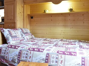 Ein Interieur einer Holzhütte mit einem Bett, das eine geblümte Patchworkdecke zeigt, einer Holzkommode und einem Regal mit zwei kleinen Behältern.