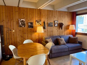 Ein gemütliches Zimmer mit einer Holzwandverkleidung, einem grauen Sofa, einem runden Holztisch mit weißen Stühlen und einem Fenster mit Vorhängen.