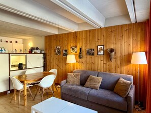 A cozy living room with a wooden wall, a grey sofa, a dining table with chairs, and a white coffee table. - Apartment with balcony - 4 guests, forum view (Flaine)