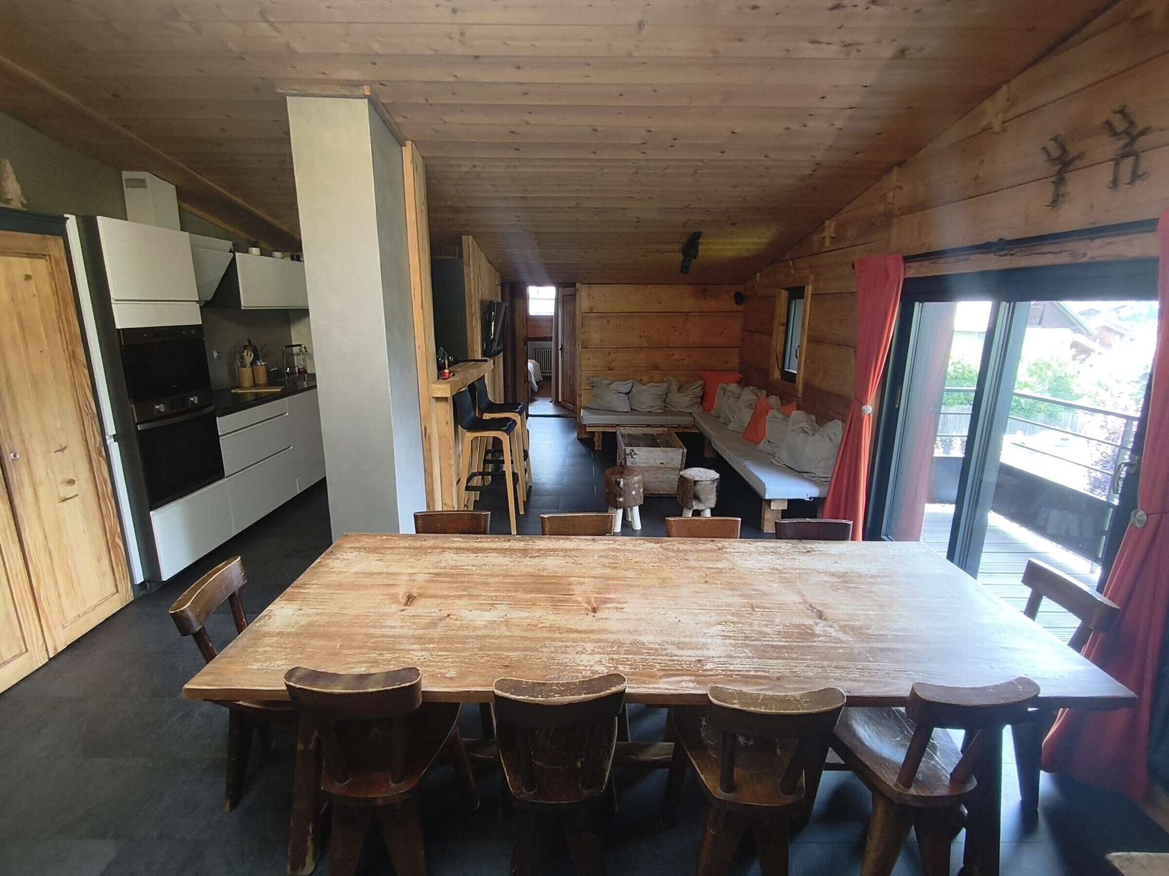 Een houten interieur met een eettafel en stoelen, een keuken met apparatuur, en uitzicht door een raam.