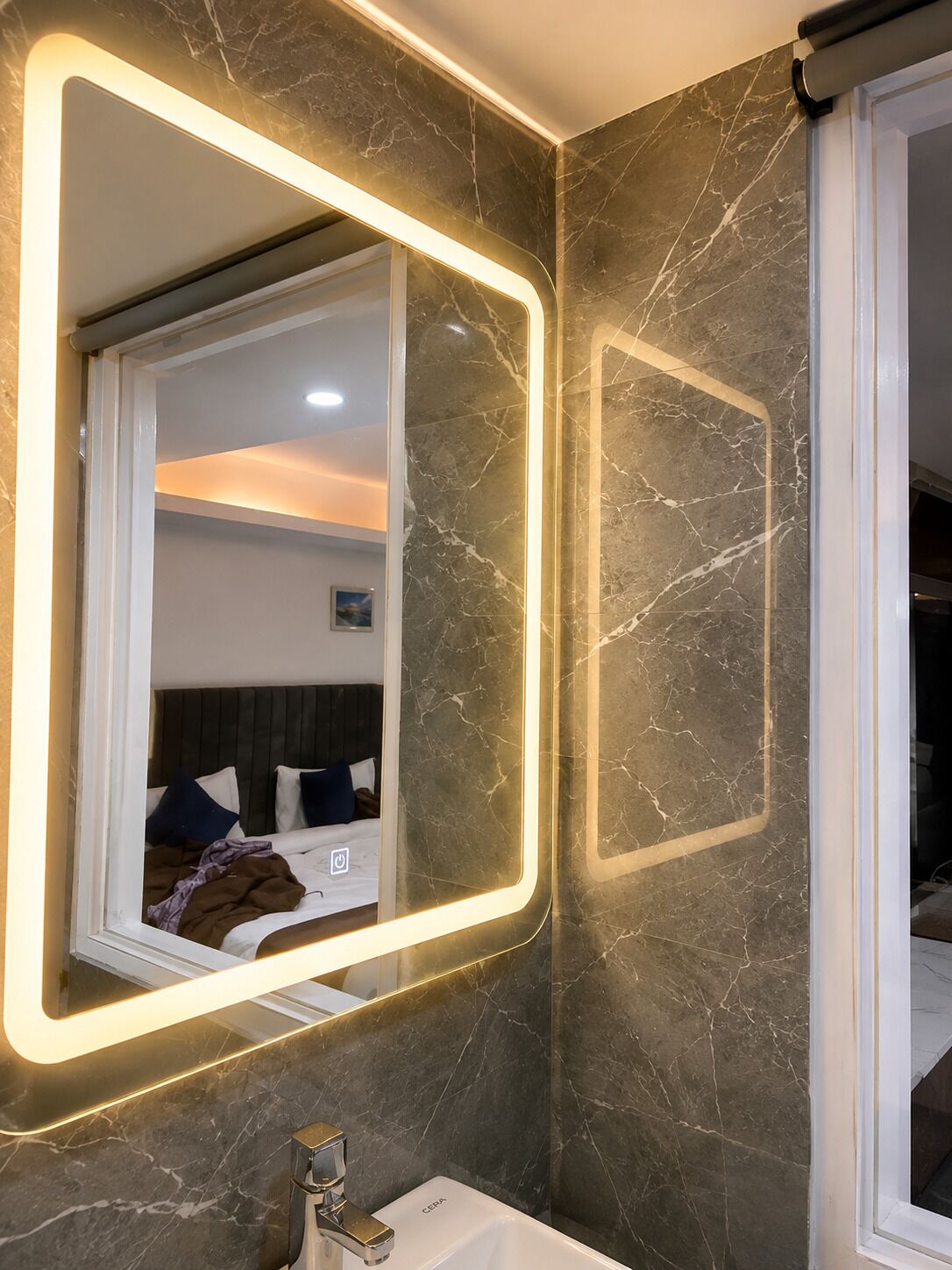 Un baño moderno con un espejo grande, paredes de mármol y una cama con almohadas a lo lejos.