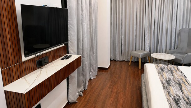 A modern hotel room with a flat-screen TV mounted on a wooden panel, a small table with a chair, a sofa, and a large bed with a patterned bedspread. - PRISHA HOTAL AND RESTAURANTS (Zirakpur)