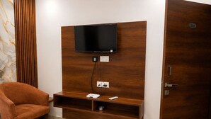 A room with a wooden entertainment unit, a television mounted above it, a chair, and a door with a number plate. - PRISHA HOTAL AND RESTAURANTS (Zirakpur)