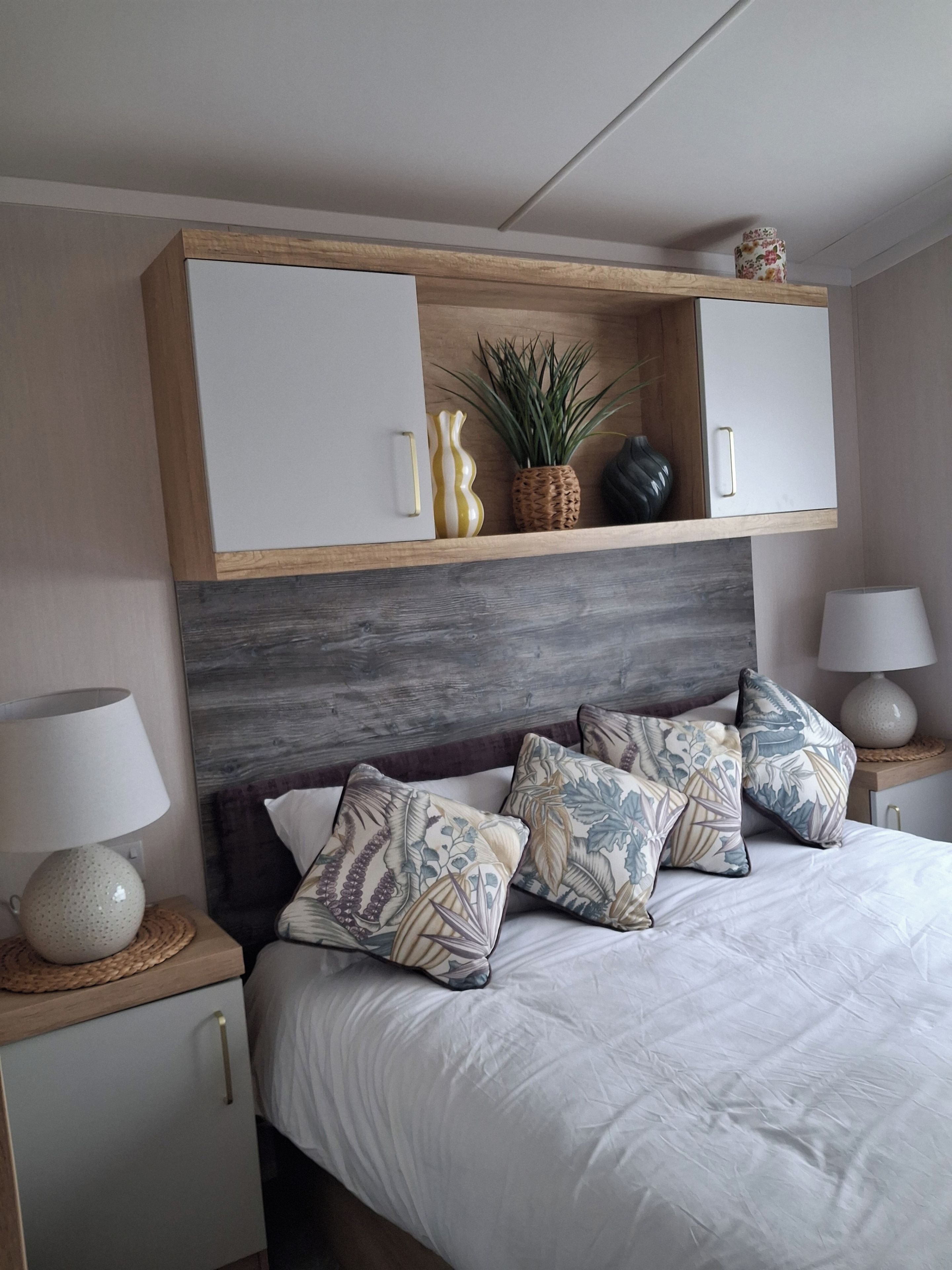 A bedroom with a bed, two bedside tables, a nightstand, a wooden headboard, a shelf with decorative items, and a wall-mounted cabinet.