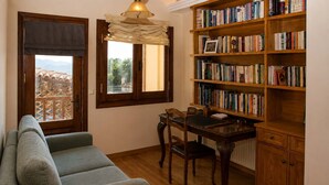 A cozy living room with a sofa, a dining table with chairs, a bookshelf, and a window with curtains. - Mountain Breeze Sanctuary with View near Volos (Alli Meria)