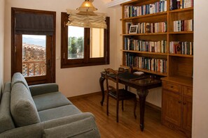 A cozy living room with a sofa, a dining table with chairs, a bookshelf, and a window with curtains. - Mountain Breeze Sanctuary with View near Volos (Alli Meria)