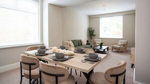 En matplats med ett bord dukat för sex personer, omgivet av sex stolar, i ett modernt vardagsrum med en soffa och ett soffbord.