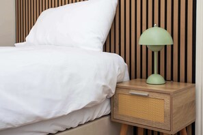 Una cama con ropa blanca, una mesita de noche de madera con una lámpara verde y una cesta tejida.
