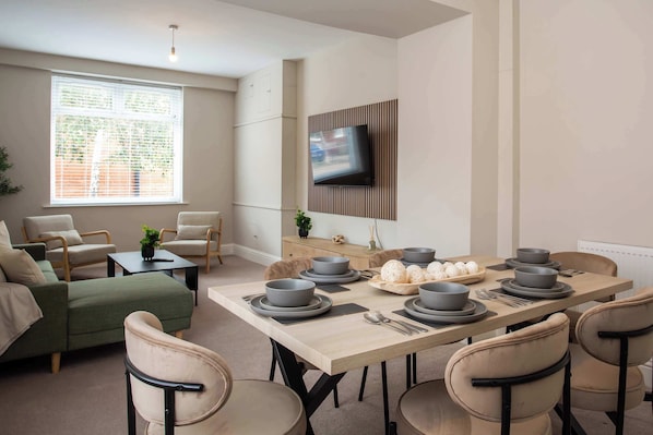 A modern living room with a dining area, a flat-screen TV mounted on a wooden panel, and a large window with blinds.