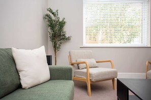 Una habitación con un sofá verde, una silla de madera, una mesita y una ventana con persianas.