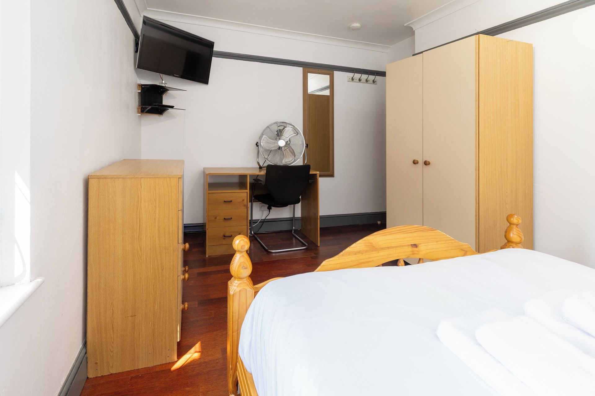 A bedroom with a bed, a desk, a chair, a wardrobe, and a television mounted on the wall.