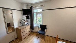 Ein Zimmer mit einem Holzkapuzenbett, einem schwarzen Stuhl, einem Standventilator, einem an der Wand befestigten Fernseher und einem Fenster mit Blick auf Grünflächen.