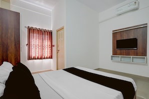 A hotel room with a bed, a TV mounted on the wall, a window with curtains, and an air conditioning unit.