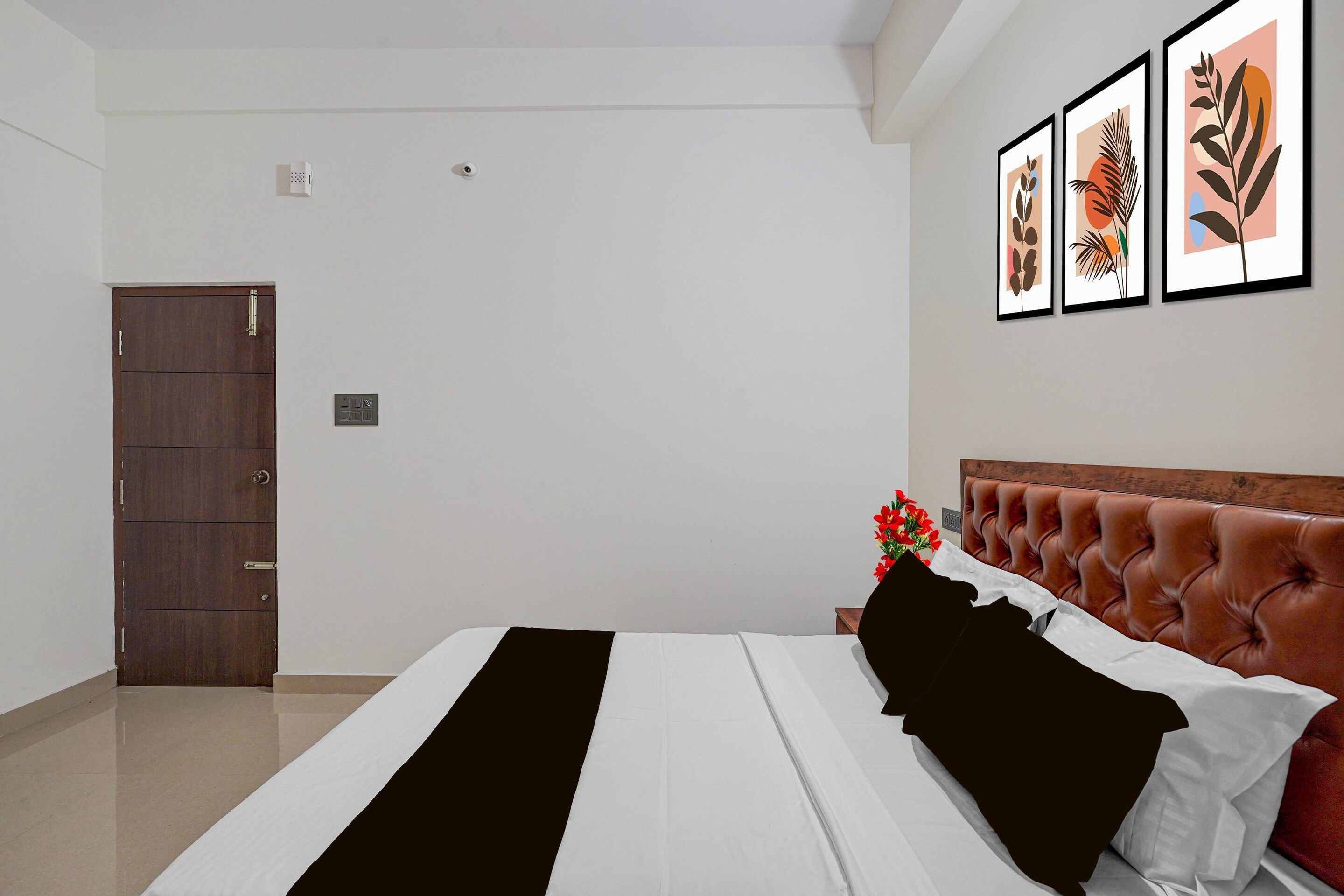 A bedroom with a bed, a wooden wardrobe, a nightstand with a vase of flowers, and three framed artworks on the wall.
