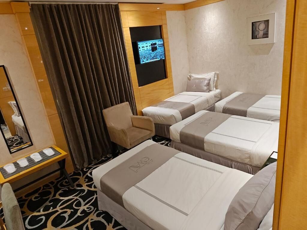 A hotel room with three beds, a TV mounted on the wall, a small sofa, and a mirror with a towel rack.