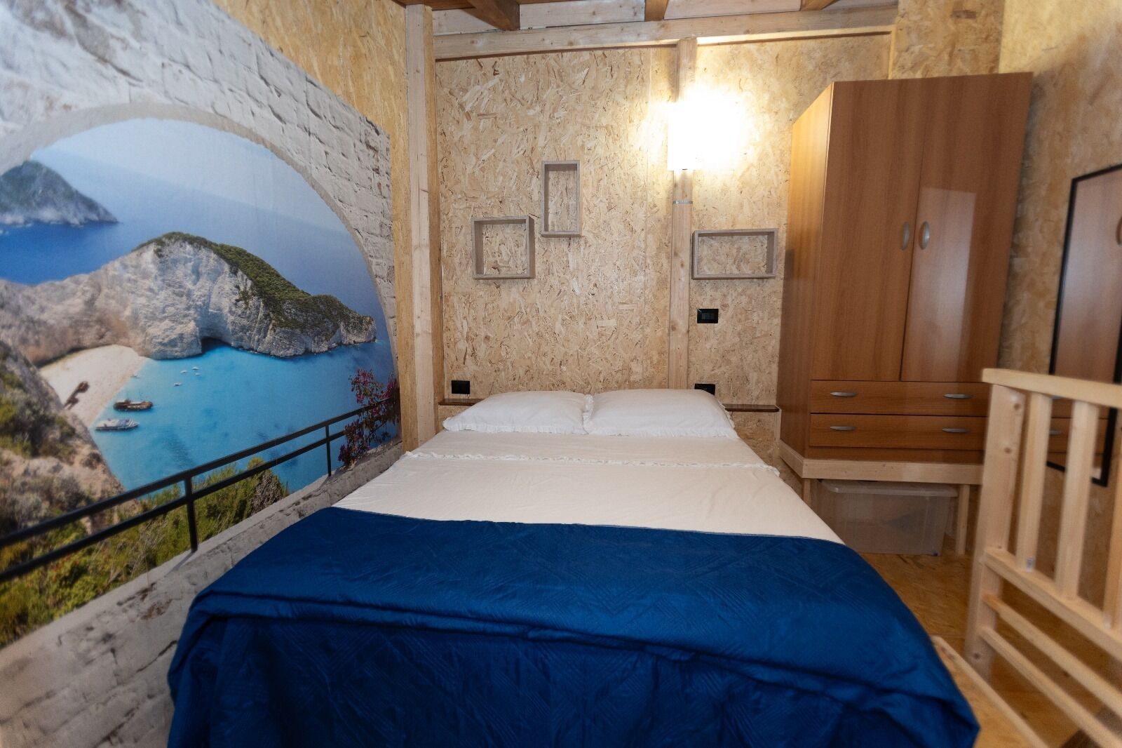 A bedroom with a bed, wooden wardrobe, and a mural of a coastal scene.