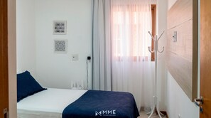 Ein Hotelzimmer mit Bett, Kleiderhaken und einem Fenster mit durchsichtigen Vorhängen.