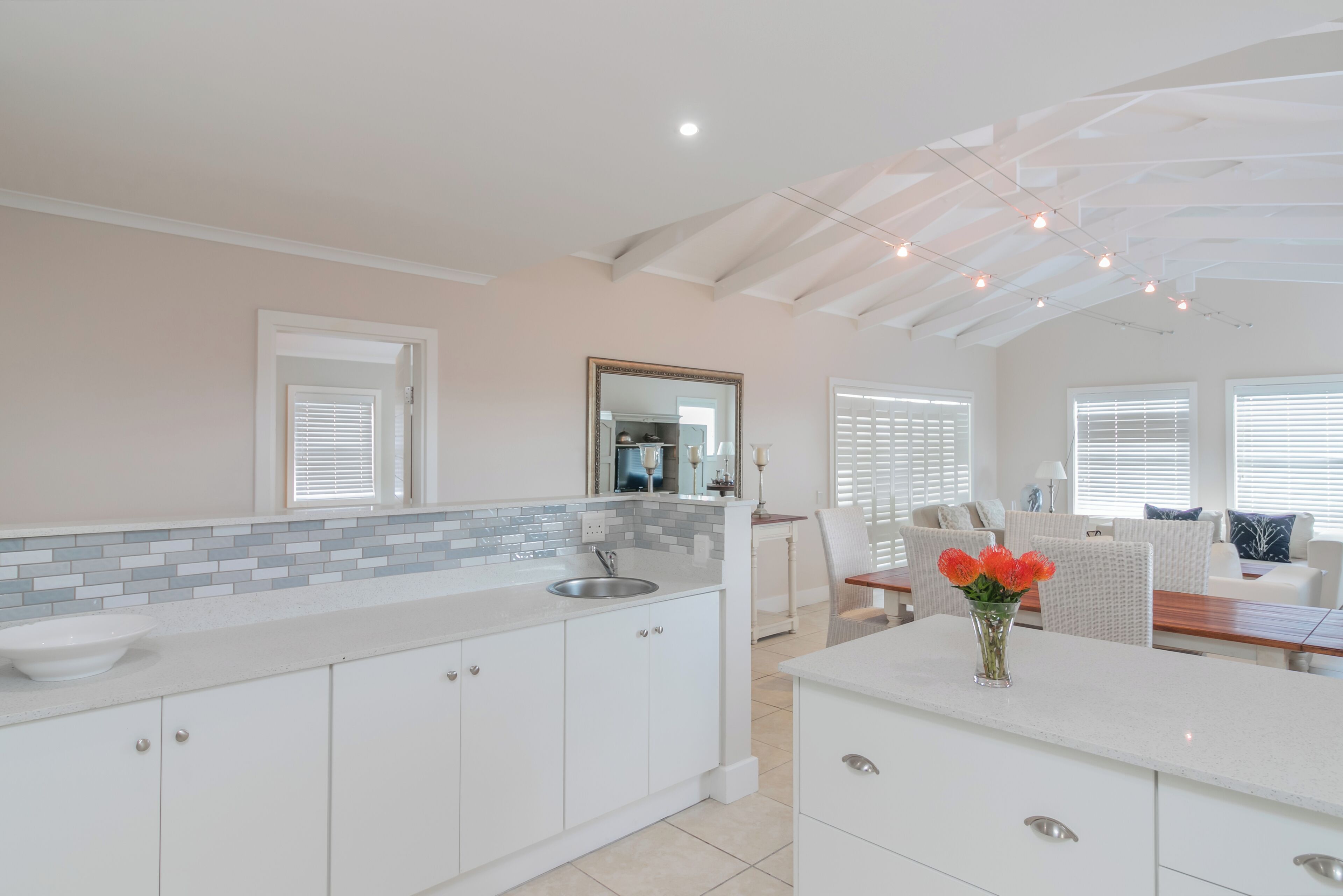 A modern kitchen with white cabinetry, a tiled backsplash, and a central island with a sink. There is a dining area with a table and chairs, and a large mirror above the island.