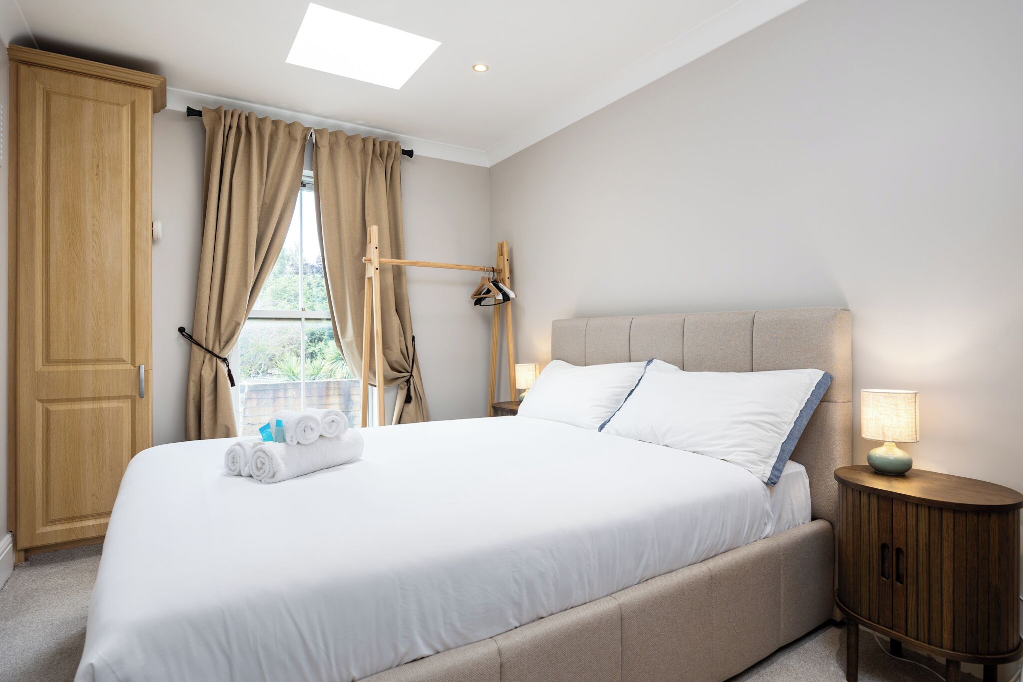 A neatly made bed with white linens, a wooden wardrobe, a bedside table with a lamp, and a window with curtains.