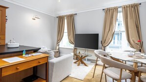 Et moderne hotelværelse med et træskrivebord, et fladskærms-tv, et spisebord med stole og store vinduer med gardiner.