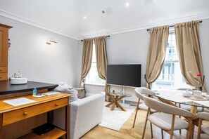 Ein modernes Hotelzimmer mit Schreibtisch aus Holz, einem Flachbildfernseher, einem Esstisch mit Stühlen und großen Fenstern mit Vorhängen.