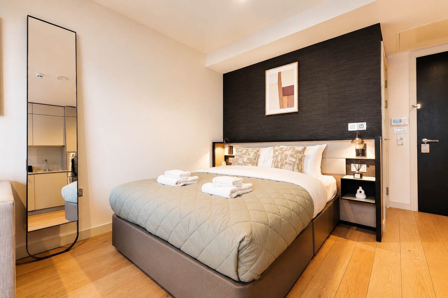A hotel room with a bed, bedside table, a mirror, a dark wall with a picture, and a door.