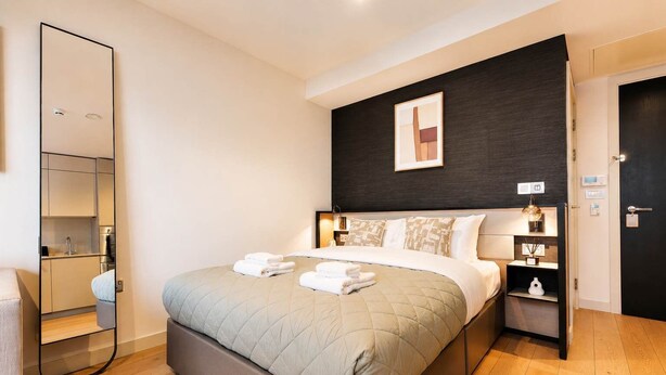 Habitación de hotel con cama, mesita de noche, espejo, pared oscura con un cuadro y una puerta.