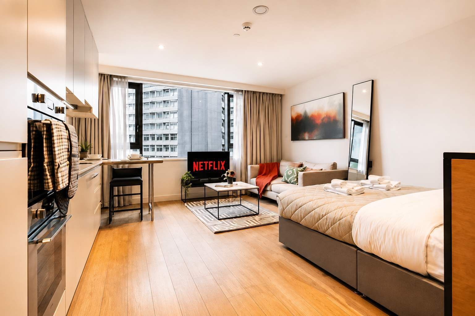 A modern hotel room with a bed, a desk with a laptop, a TV, and a view of the city.