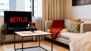 Una sala moderna con una televisión de pantalla plana mostrando 'NETFLIX', un sofá con una manta roja, un cojín estampado y una mesita con un libro y una planta.