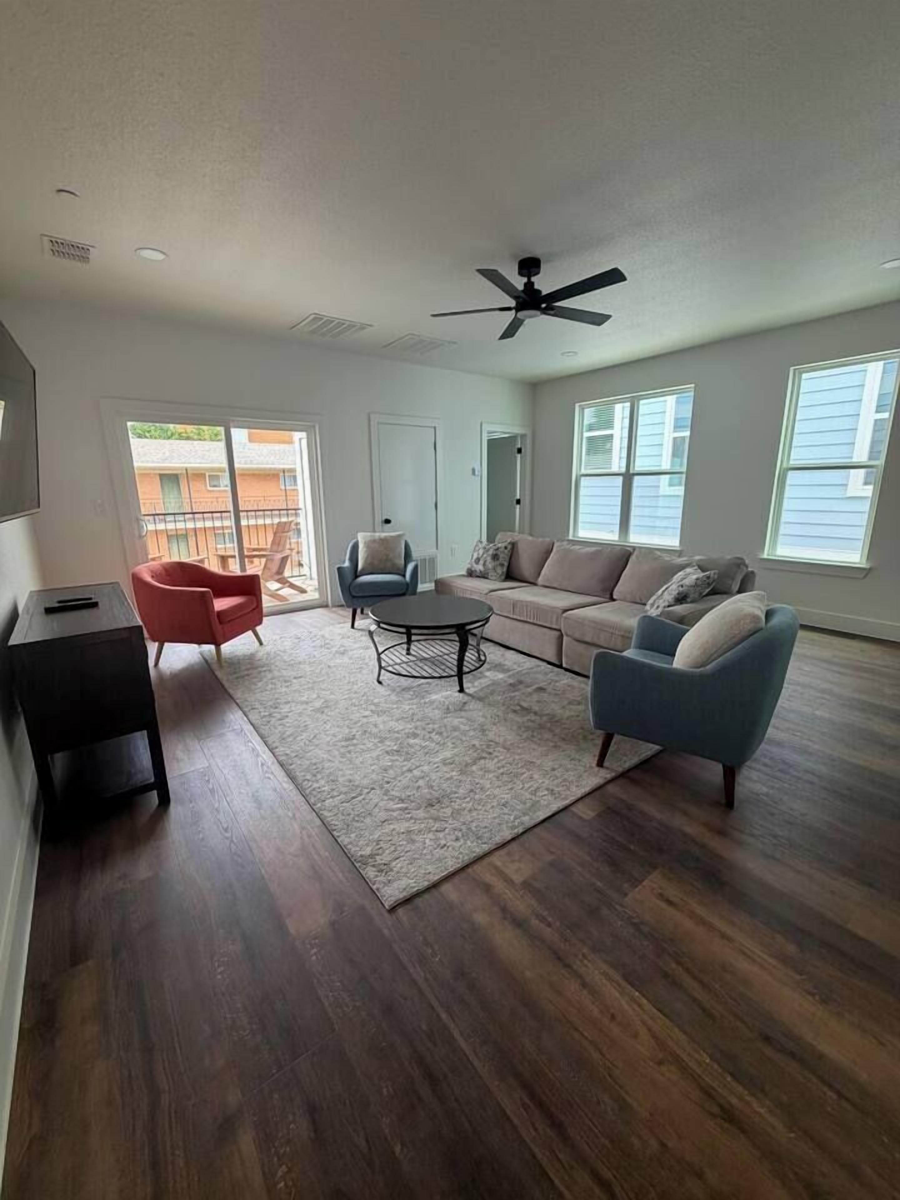 A spacious living room with a dark wood floor, a ceiling fan, a sofa, a coffee table, and a red armchair.