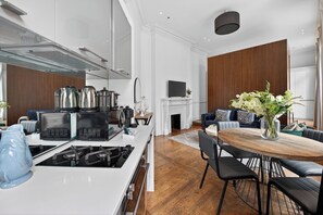 A modern kitchen with stainless steel appliances, a dining area with a wooden table and black chairs, and a living room with a fireplace and a television. - VESTO Victoria (London)