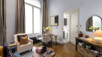 Sala de estar bem iluminada com sofá, mesa de centro e área de jantar com cadeiras. Há uma lareira, um espelho e uma janela com cortinas.