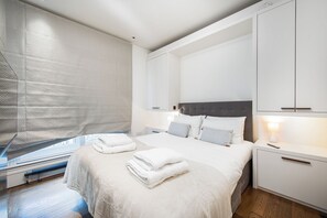 A modern bedroom with a large bed, a bedside table with a lamp, and a window with a view of the city. - VESTO Victoria (London)