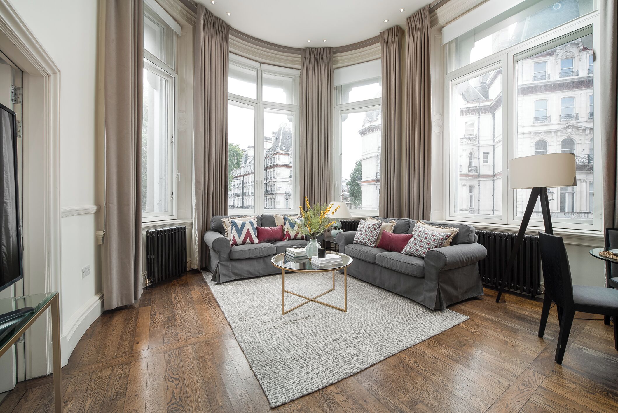 Uma sala de estar moderna com um sofá cinzento, uma mesa de centro e vista para edifícios da cidade através de grandes janelas.