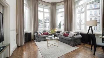 Uma sala de estar moderna com um sofá cinzento, uma mesa de centro e vista para edifícios da cidade através de grandes janelas.