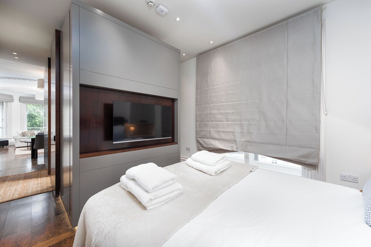 Ein modernes Schlafzimmer mit einem großen Bett, einem Flachbildfernseher an der Wand und einem großen Fenster mit Blick ins Grüne.