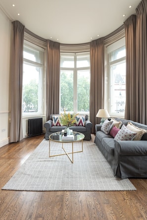 Una sala de estar moderna con un sofá gris, una mesa de centro de vidrio y vistas a la ciudad a través de grandes ventanales.
