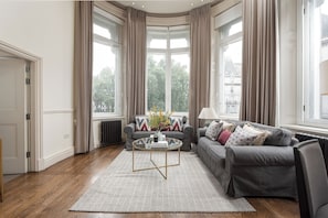 Ein modernes Wohnzimmer mit einem grauen Sofa, einem runden Couchtisch und Blick auf eine Stadtlandschaft durch große Fenster.