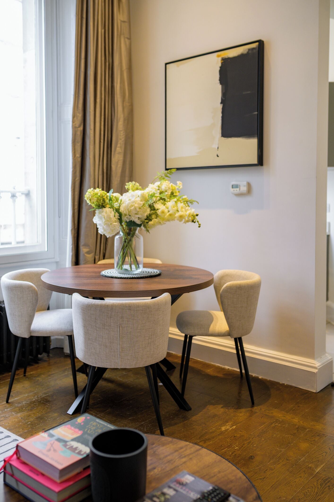 A dining area with a round table, four chairs, a vase with flowers, and a painting on the wall.