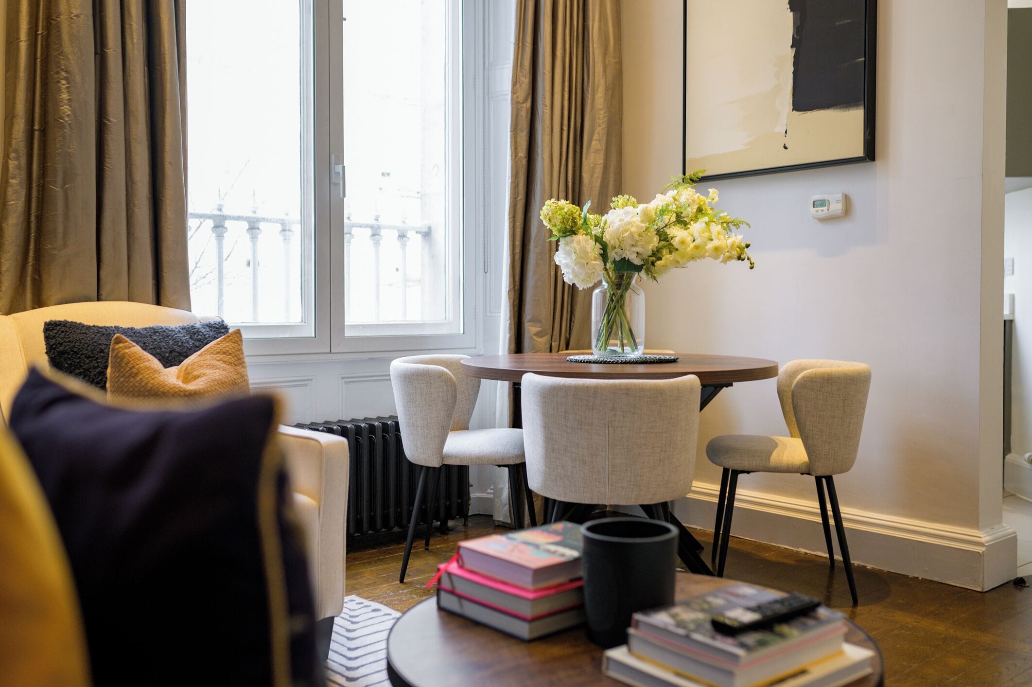 A living room with a round table, four chairs, a vase with flowers, and a bookshelf with books.