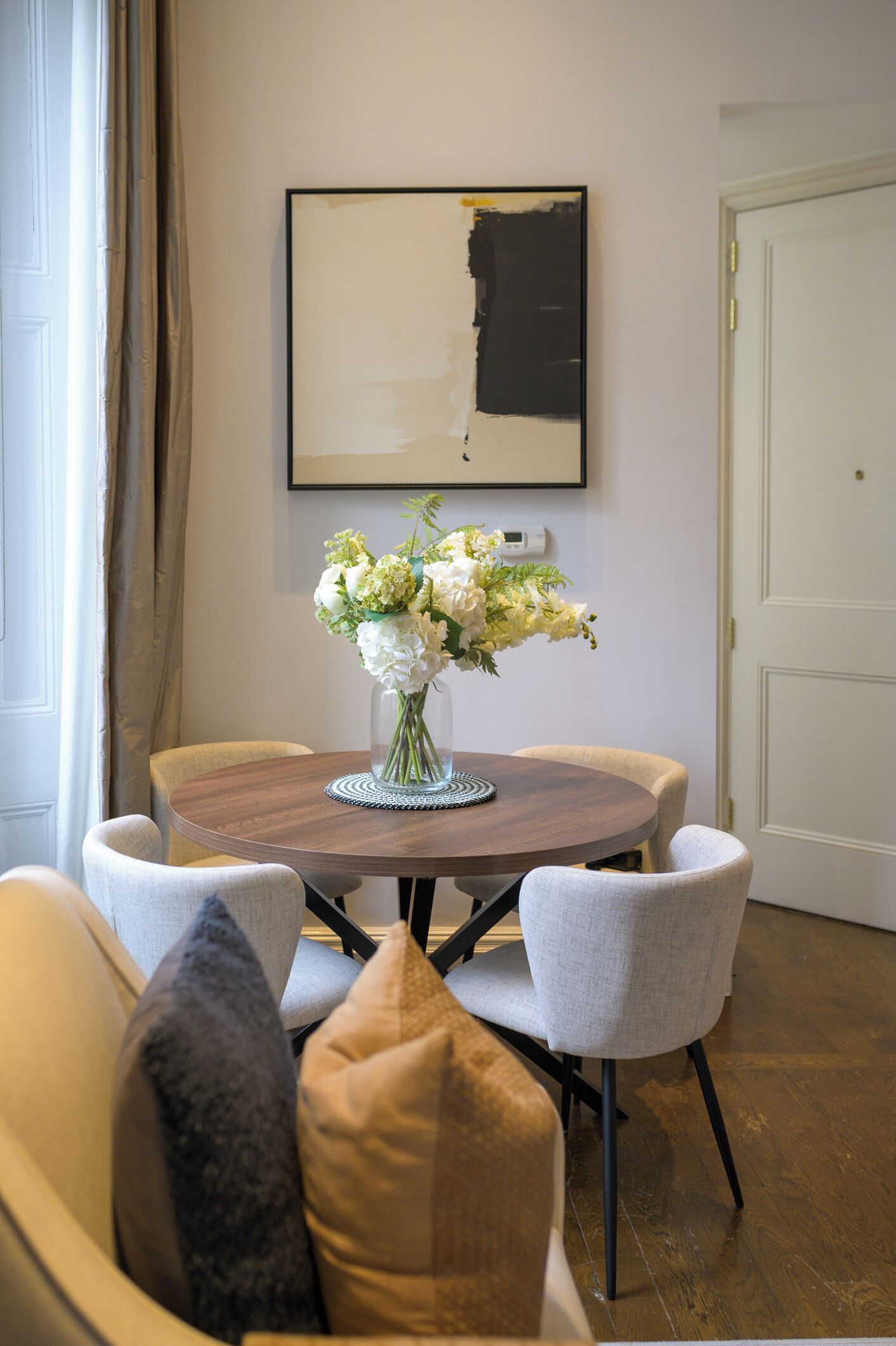 Uma sala de jantar com uma mesa redonda, cadeiras, um vaso com flores e um quadro na parede.